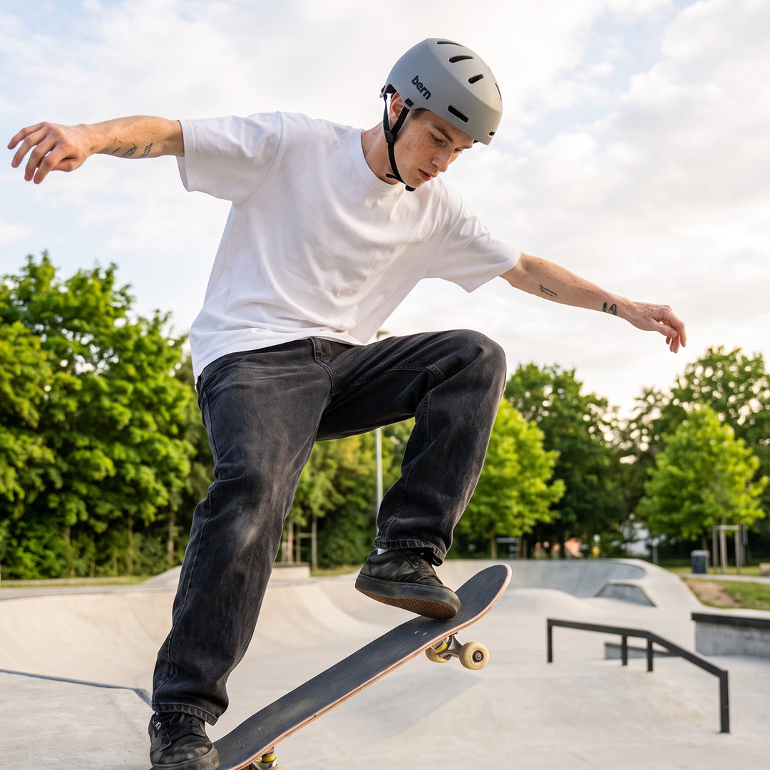 A skateboarder in a white t-shirt, dark jeans, and a gray macon 2.0 bern helmet performs a trick on a concrete skatepark ramp with trees in the background.